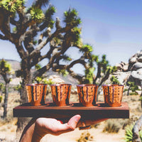 Hammered copper shot glasses on wooden board with Joshua tree in background