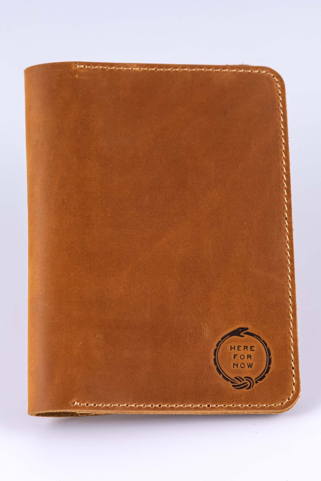 Brown leather field notes wallet closed to show the front embossed with the "Here For Now" Ouroboros design and light colored tan stitching
