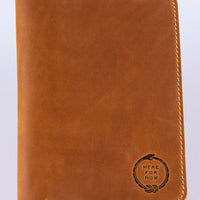 Brown leather field notes wallet closed to show the front embossed with the "Here For Now" Ouroboros design and light colored tan stitching