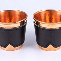 Two hammered copper cups with black leather sleeves stamped with Sound As Ever Logo on the front with black stitching