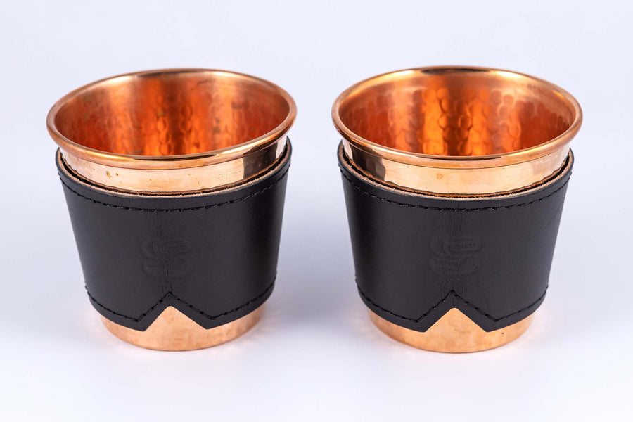 Two hammered copper cups with black leather sleeves stamped with Sound As Ever Logo on the front with black stitching