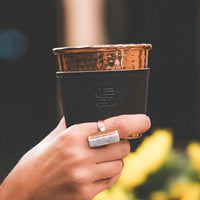 Hammered copper cup with black leather sleeve stamped with Sound As Ever Logo held up by a hand with a copper and white ring