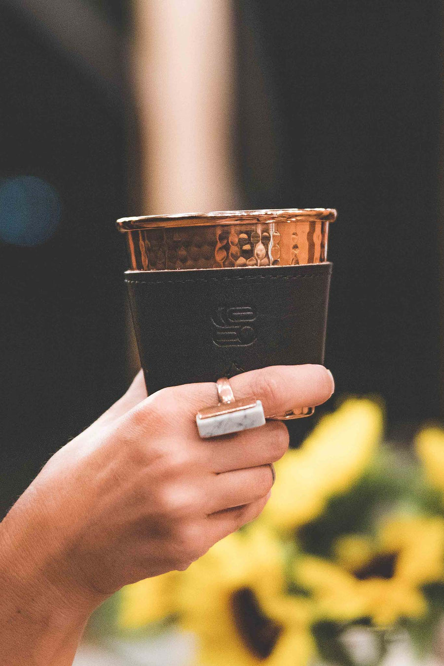 Hammered copper cup with black leather sleeve stamped with Sound As Ever Logo held up by a hand with a copper and white ring