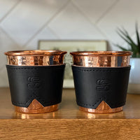 Two hammered copper cups with black leather sleeves stamped with Sound As Ever Logo on a wooden shelf 