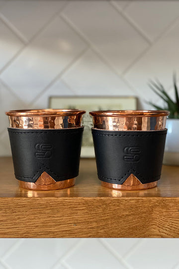 Two hammered copper cups with black leather sleeves stamped with Sound As Ever Logo on a wooden shelf 