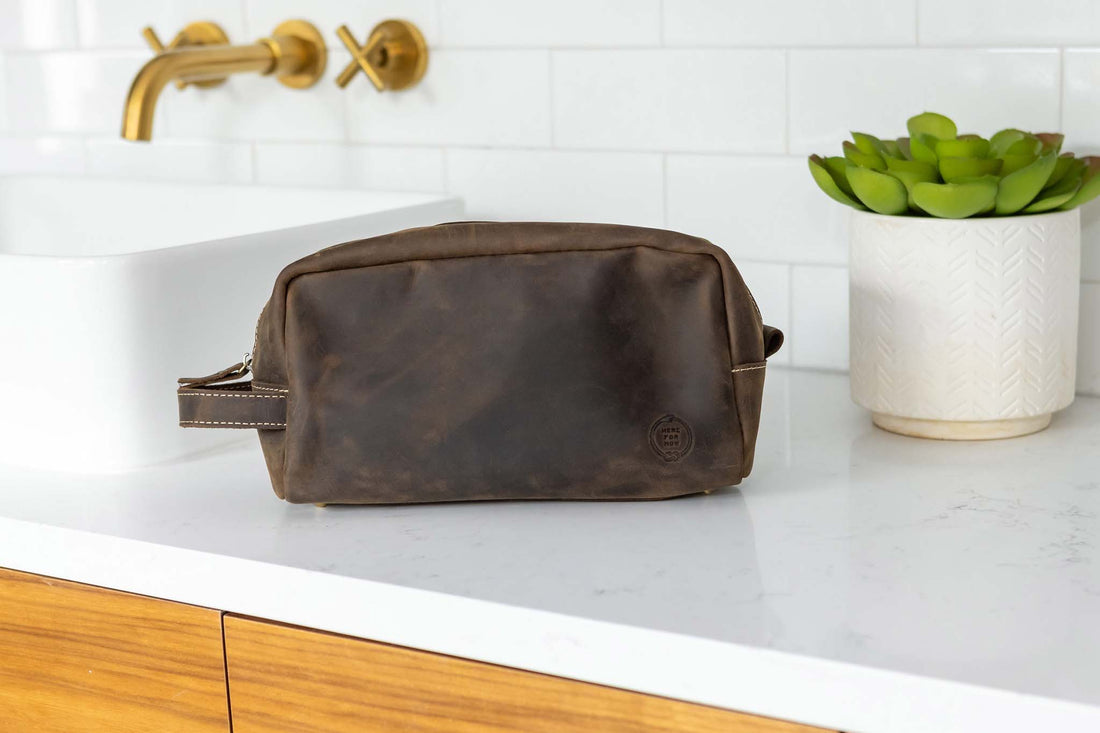 Close up view of Leather dopp bag from Sound As Ever in dark brown umber color with the embossed "Here For Now" Ouroboros design and zip closure and off-white stitching and side strap handle