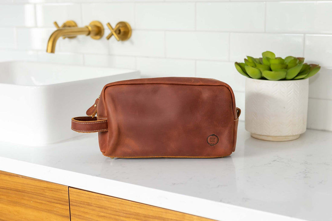 Close up view of Leather dopp bag from Sound As Ever in rich reddish brown rust color with the embossed "Here For Now" Ouroboros design and zip closure and side strap handle