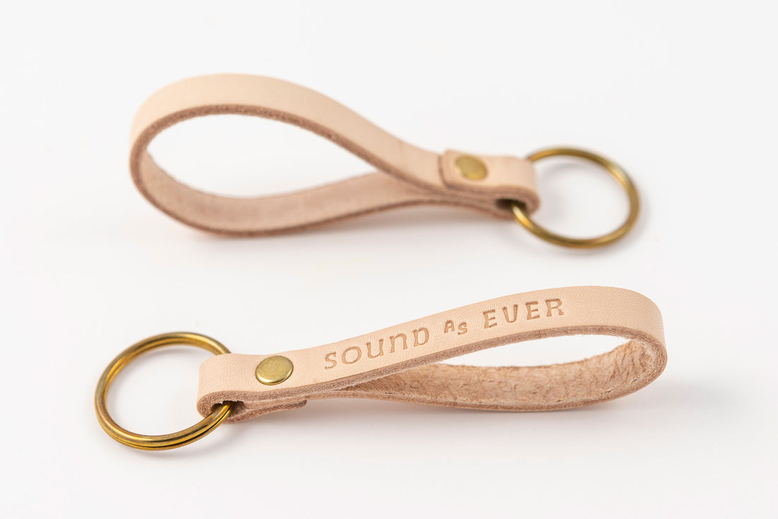 Landscape orientation view of tan color leather keychain with brass ring stamped with text "Sound as Ever"