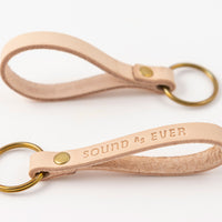Landscape orientation view of tan color leather keychain with brass ring stamped with text "Sound as Ever"