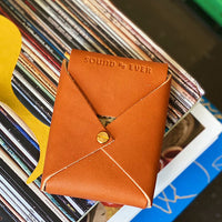 Leather playing card case on top of books clearly displaying stamped letters "Sound As Ever" on opening flap