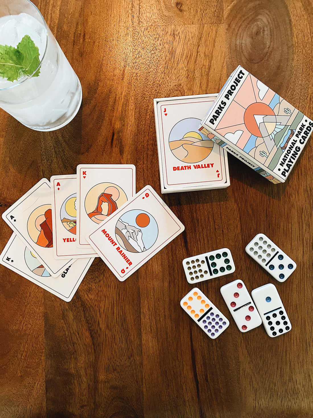 National park playing cards splayed on a table with their box, some dominos and a drink with a mint leaf