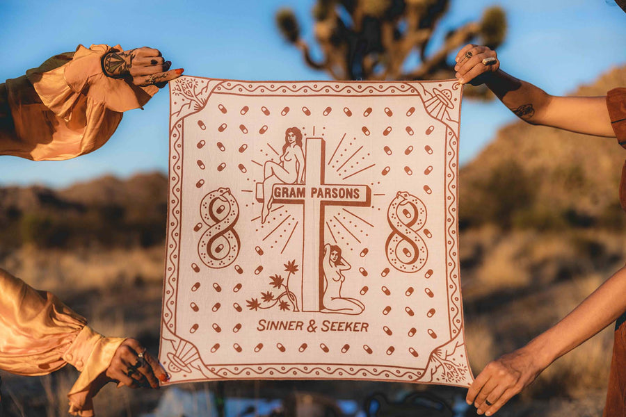 Two women holding the Sinner & Seeker organic cotton bandana in the desert. The design has two women next to a cross, joshua trees, UFOs, and snakes in an infinity shape
