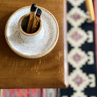 Palo santo holder and sage burning bowl made from white speckled clay with two burnt palo santo sticks  on a table with a patterned rug beneath