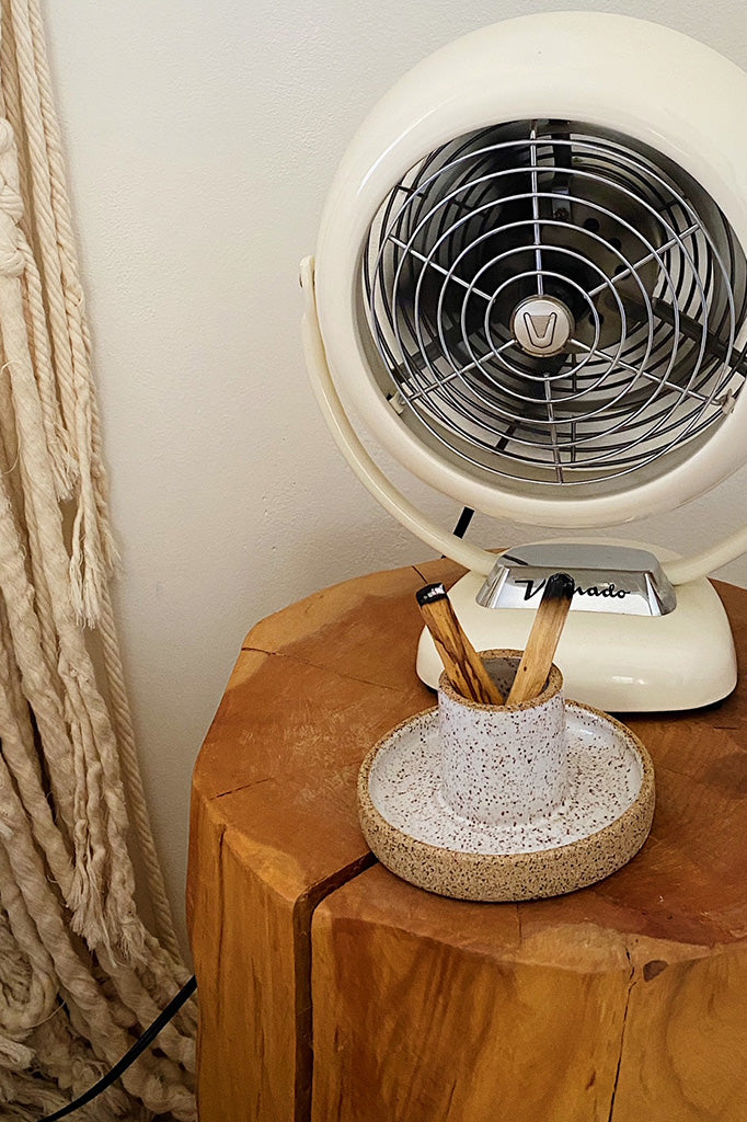 Palo santo holder and sage burning bowl made from white speckled clay with two burnt palo santo sticks next to a vintage fan on a wood side table