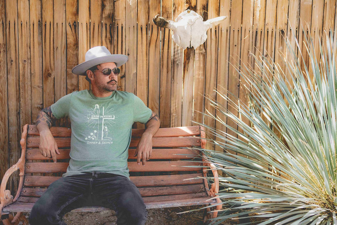 Man sitting on a wooden bench near a sotol plant wearing Sinner & Seeker green retro graphic tee and flat brim hat