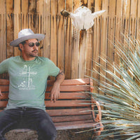 Man sitting on a wooden bench near a sotol plant wearing Sinner & Seeker green retro graphic tee and flat brim hat
