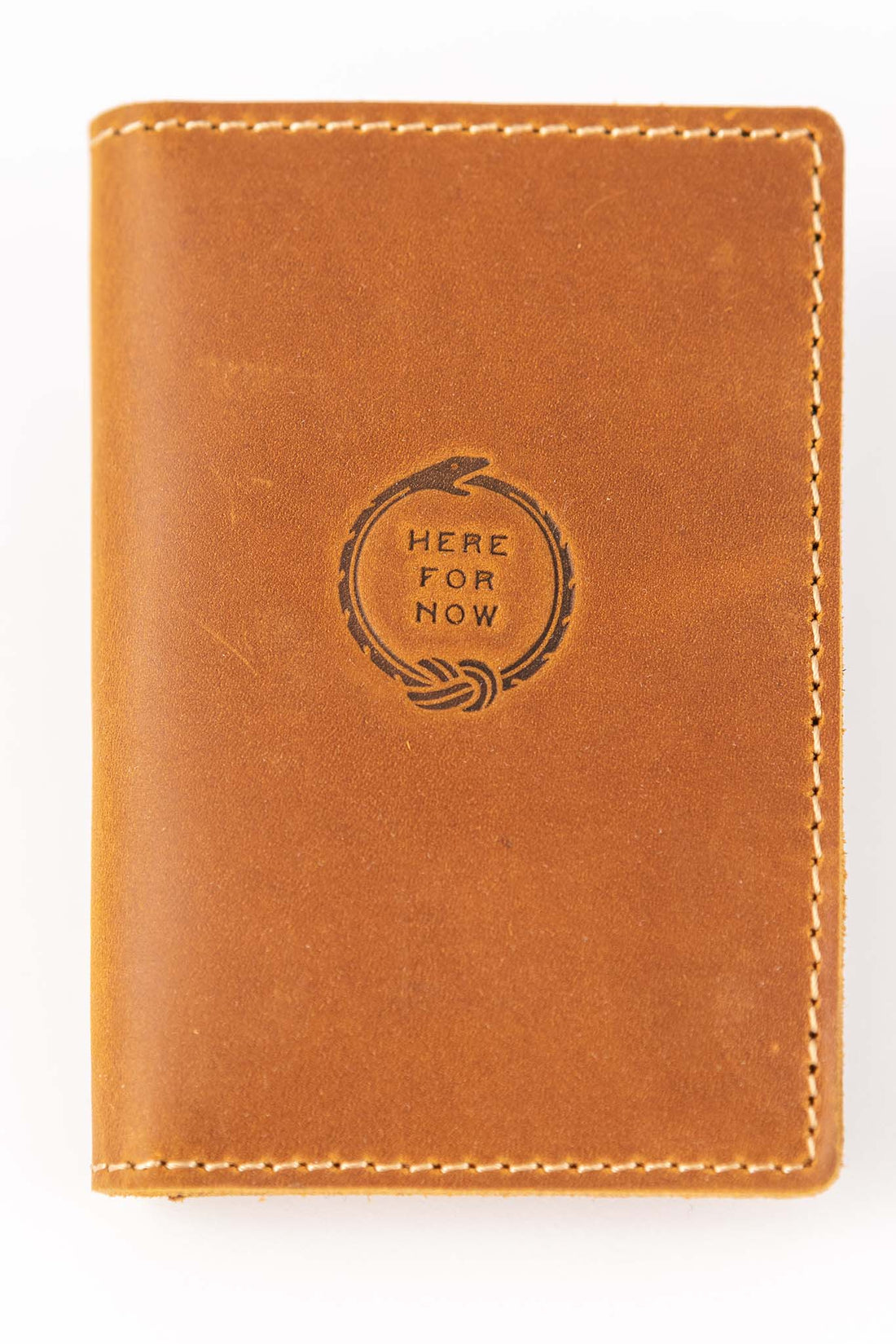 Front and closed view of slim bifold tan colored leather wallet with visible stitching and stamped Ouroboros design with text "Here For Now"