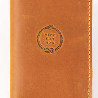 Front and closed view of slim bifold tan colored leather wallet with visible stitching and stamped Ouroboros design with text "Here For Now"
