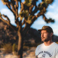 Man in desert with joshua trees in background wearing loden green Sound As Ever snapback cap and white shirt