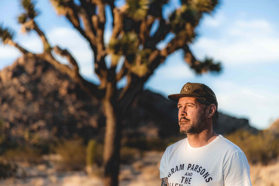 Man in desert with joshua trees in background wearing loden green Sound As Ever snapback cap and white shirt