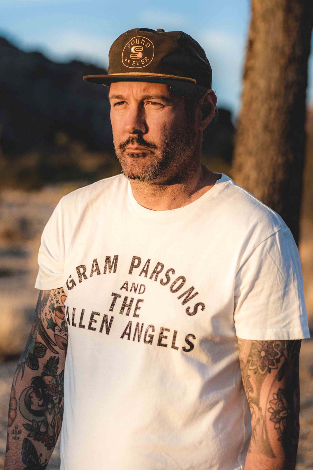 Man wearing Sound As Ever snapback hat with yellow logo and olive green material and white Gram Parsons and the Fallen Angels official classic t-shirt