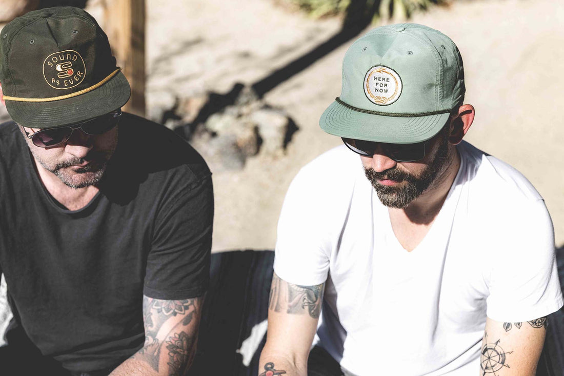 Two men sitting with one wearing Sound As Ever snapback cap and other wearing Hear For Now unstructured snapback hat