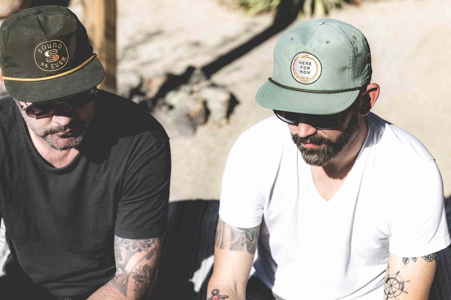 Two men sitting with one wearing Sound As Ever snapback cap and other wearing Hear For Now unstructured snapback hat