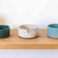 Three stoneware ashtrays on a wooden shelf in different colors. From left to right the colors are dark greenish blue, white, and light blue glaze over the speckled clay base