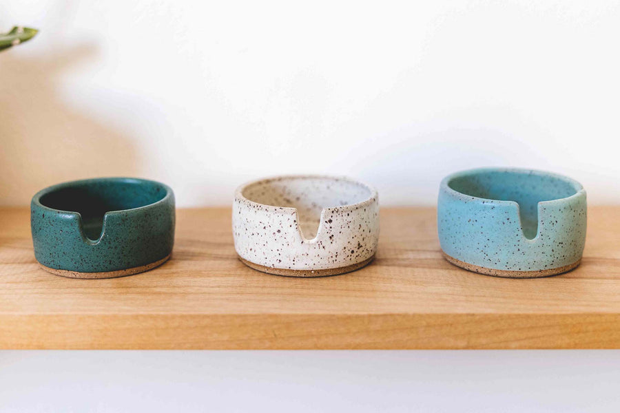 Three stoneware ashtrays on a wooden shelf in different colors. From left to right the colors are dark greenish blue, white, and light blue glaze over the speckled clay base