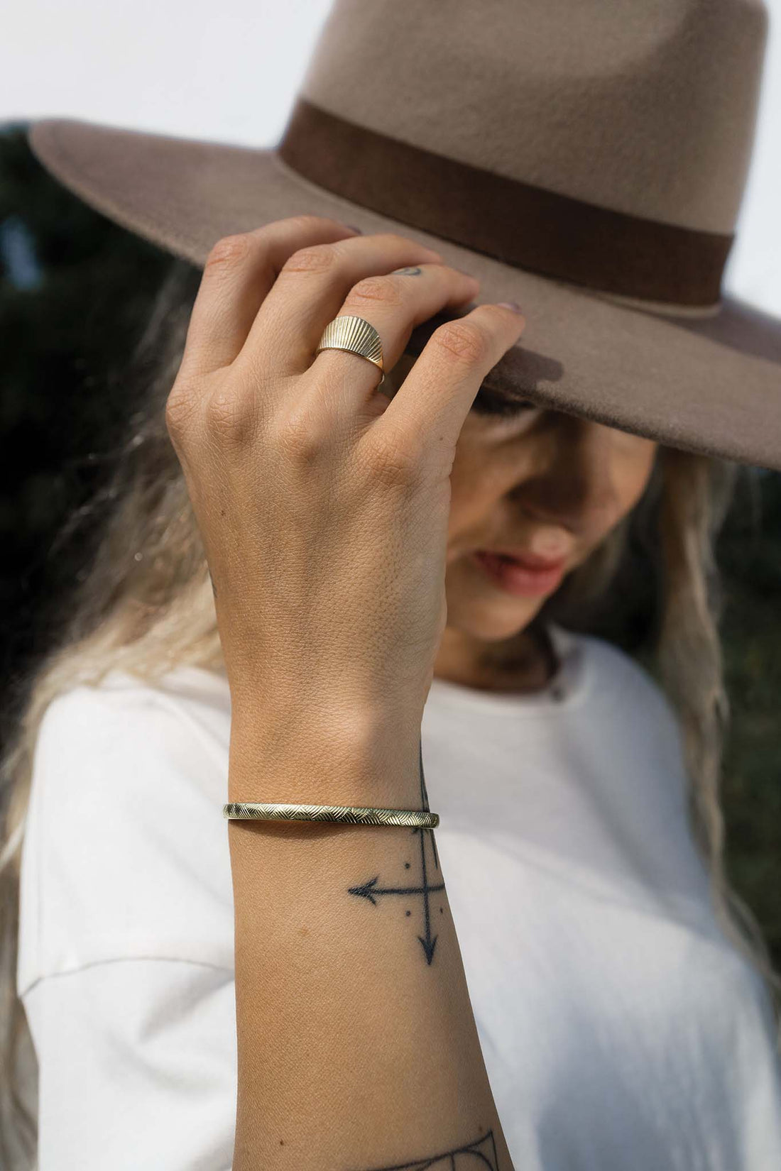 Woman's hand grabbing the side of her felt hat while wearing a brass sunburst ring and brass bracelet