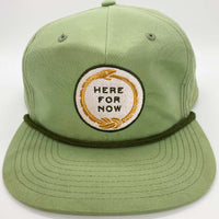 Light green unstructured snapback hat with white and gold ouroboros logo with text "Here For Now"