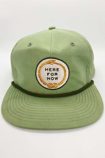 Light green unstructured snapback hat with white and gold ouroboros logo with text "Here For Now"