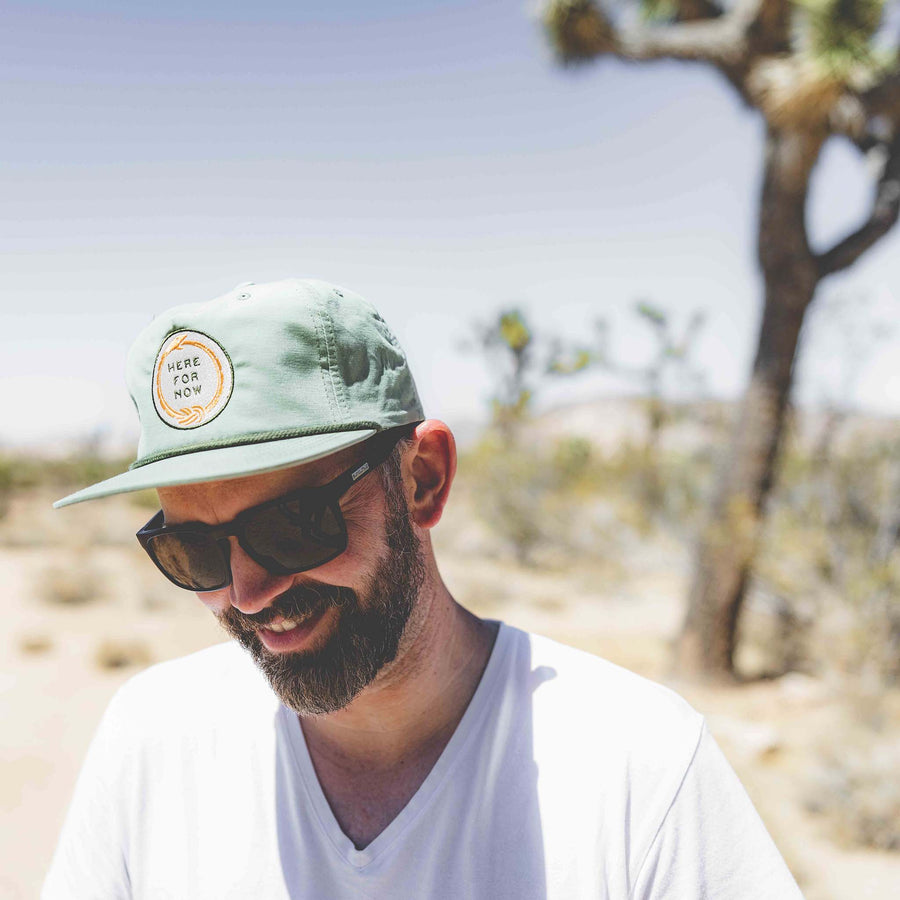 Man wearing pistachio green unstructured snapback hat with white and gold ouroboros logo with text "Here For Now"
