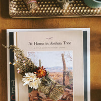 A white sage bundle decorated with desert flowers sitting on top of a book guide for desert living with the title "At Home in Joshua Tree"