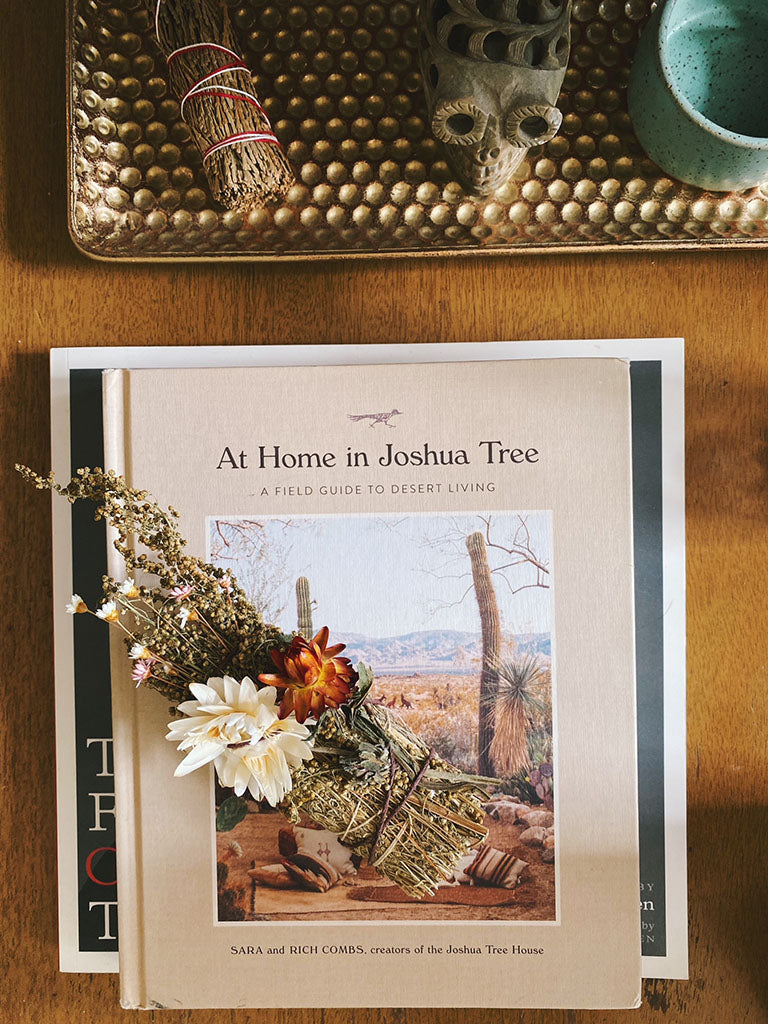 A white sage bundle decorated with desert flowers sitting on top of a book guide for desert living with the title "At Home in Joshua Tree"
