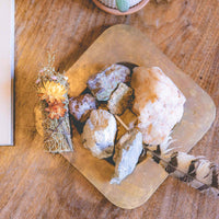 A white sage bundle decorated with desert flowers on top of a burner with rocks and large bird feather