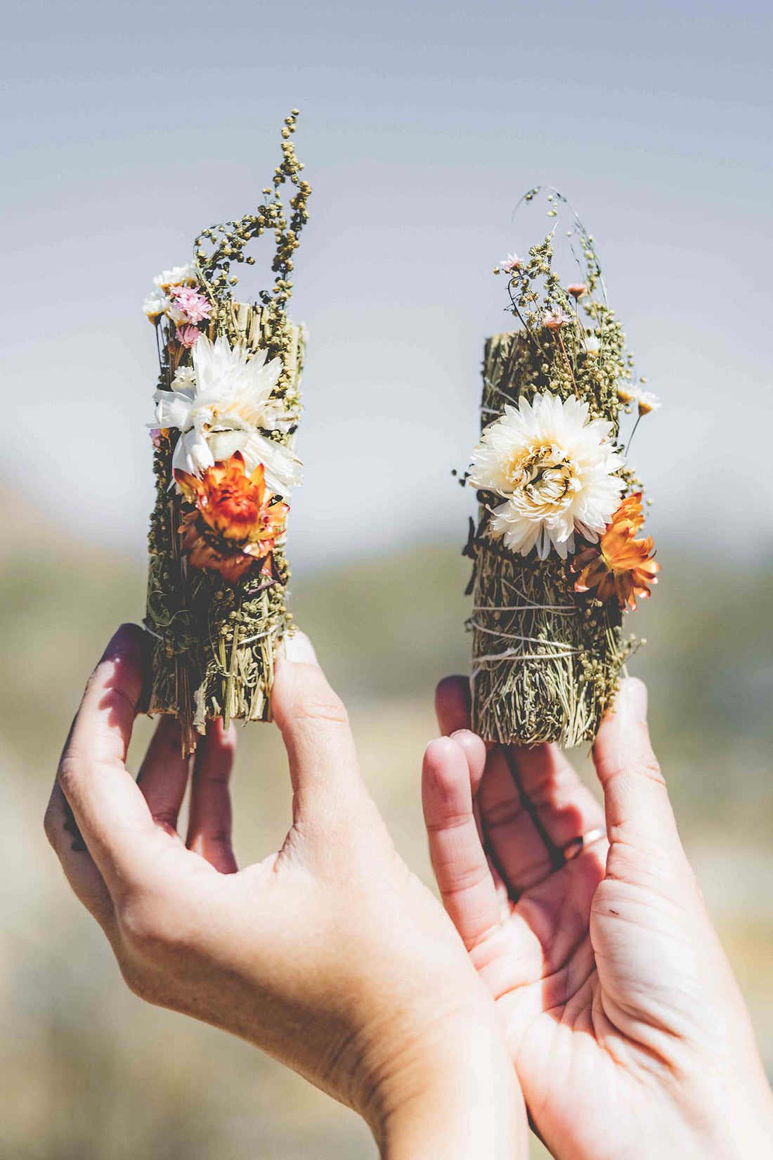 Two hands each holding a white sage bundle decorated with desert flowers