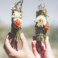 Two hands each holding a white sage bundle decorated with desert flowers