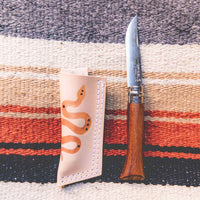 Wood handle "Whiskey" pocket knife made with an Opinel blade next to a tan leather sheath on top of an artisan wool blanket