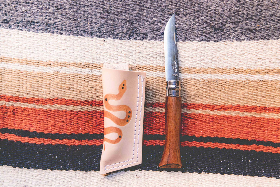 Wood handle "Whiskey" pocket knife made with an Opinel blade next to a tan leather sheath on top of an artisan wool blanket